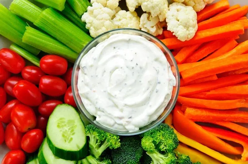 A bowl of creamy herb dip surrounded by cherry tomatoes, carrots, cucumbers, celery, broccoli, and cauliflower